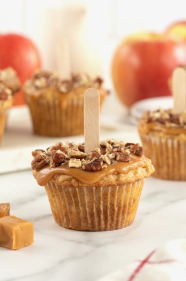 A caramel apple cupcake with caramel and pecan pieces on top and a popsicle stick in the center.