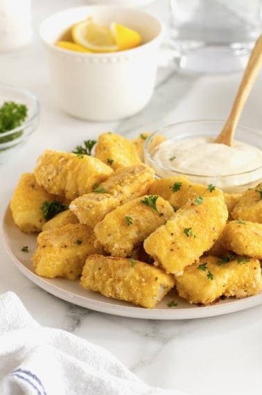 A large white serving plate with a pile of golden battered fish sticks surrounding a small glass dish of tartar sauce.