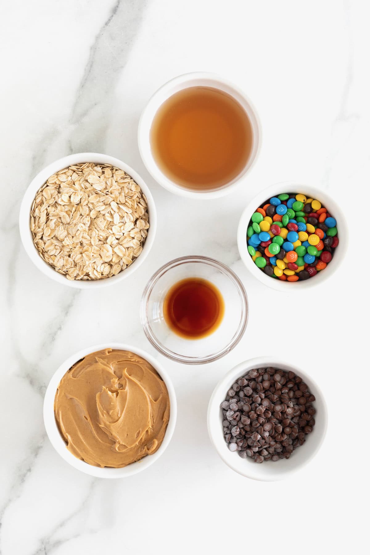Ingredients to make no-bake monster cookie balls in small white and glass dishes on a white marble counter.