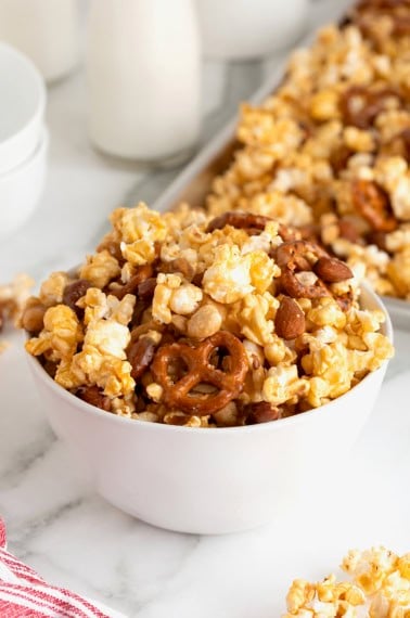 A white bowl of caramel corn mix with peanuts, pretzels and almonds on a white marble counter.