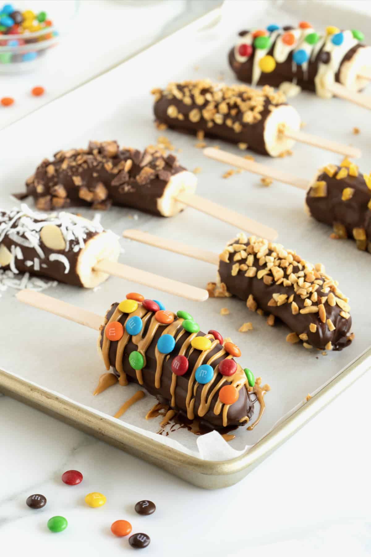 Frozen bananas with popsicles sticks covered in chocolate and mini M&Ms, mini chocolate chips, crushed peanuts and coconut.