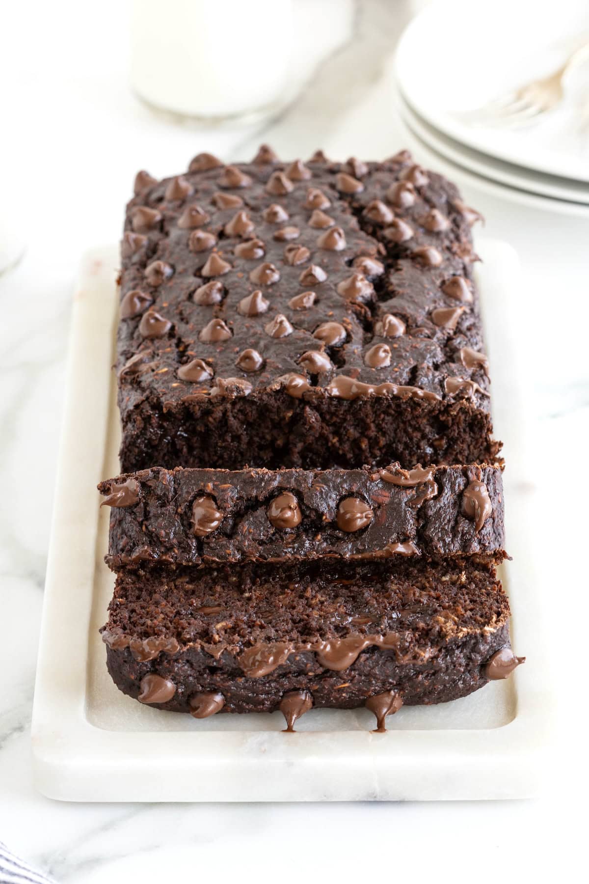 A loaf of chocolate banana bread studded with chocolate chips on top. The loaf is sitting on a white cutting board on a white marble counter.