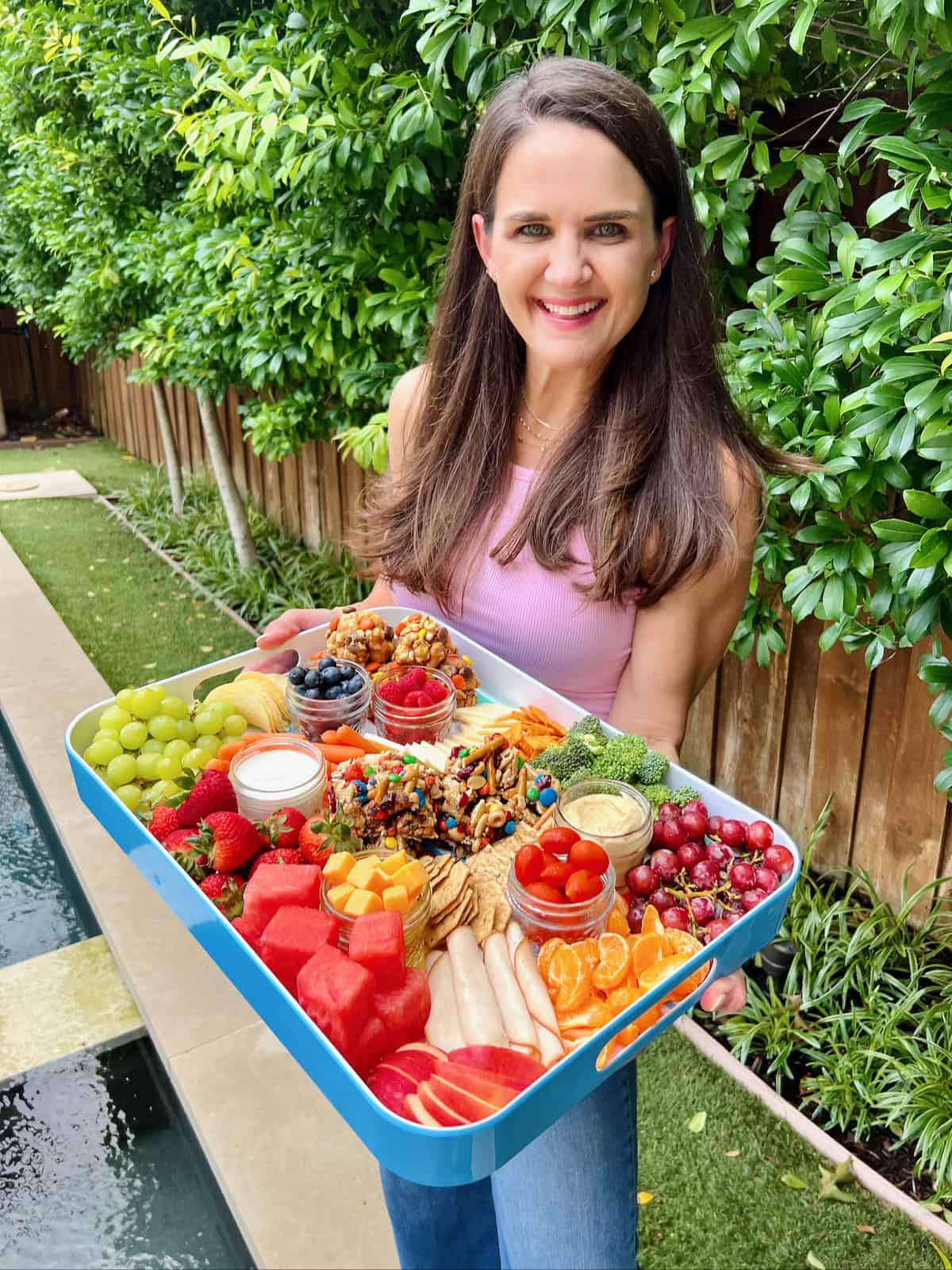 A white woman with long brown hair holding a white rimmed snack tray with handles filled with fruits, vegetables and no bake snacks.