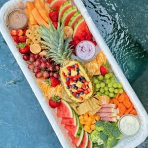 Close up shot of a snack tray floating in a pool. The tray has a halved pineapple in the center and is surrounded by fruits, cheeses and crackers.