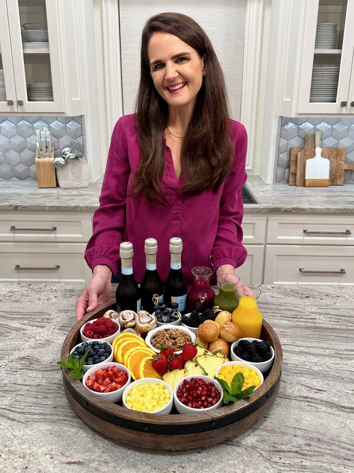 A white woman with long, dark hair wearing a magenta long sleeve shirt standing behind a mimosa board.