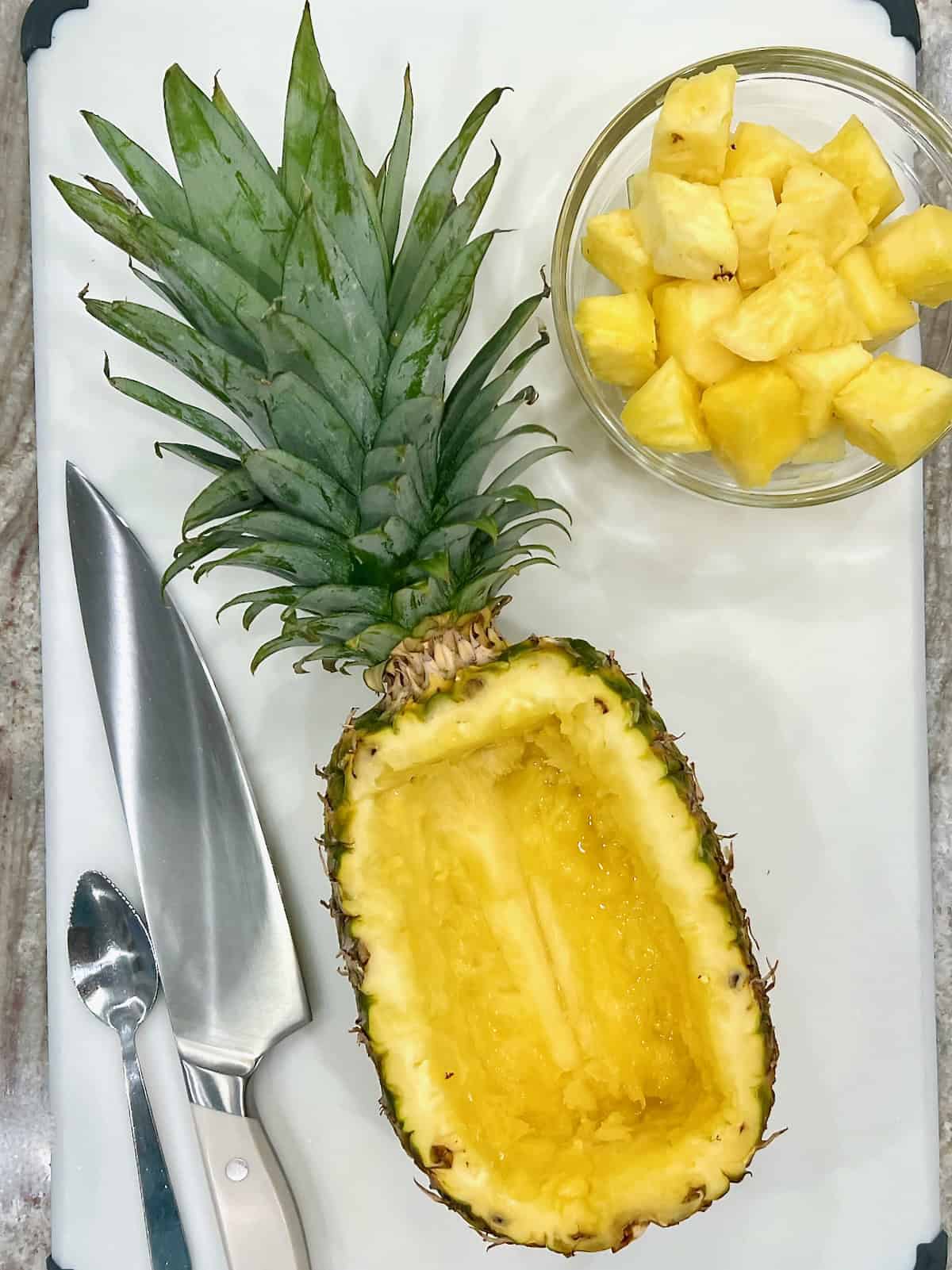 A hollowed out pineapple laying on a flat surface. A glass bowl of pineapple chunks sits to the top right and a very sharp knife lays to the lower left side.