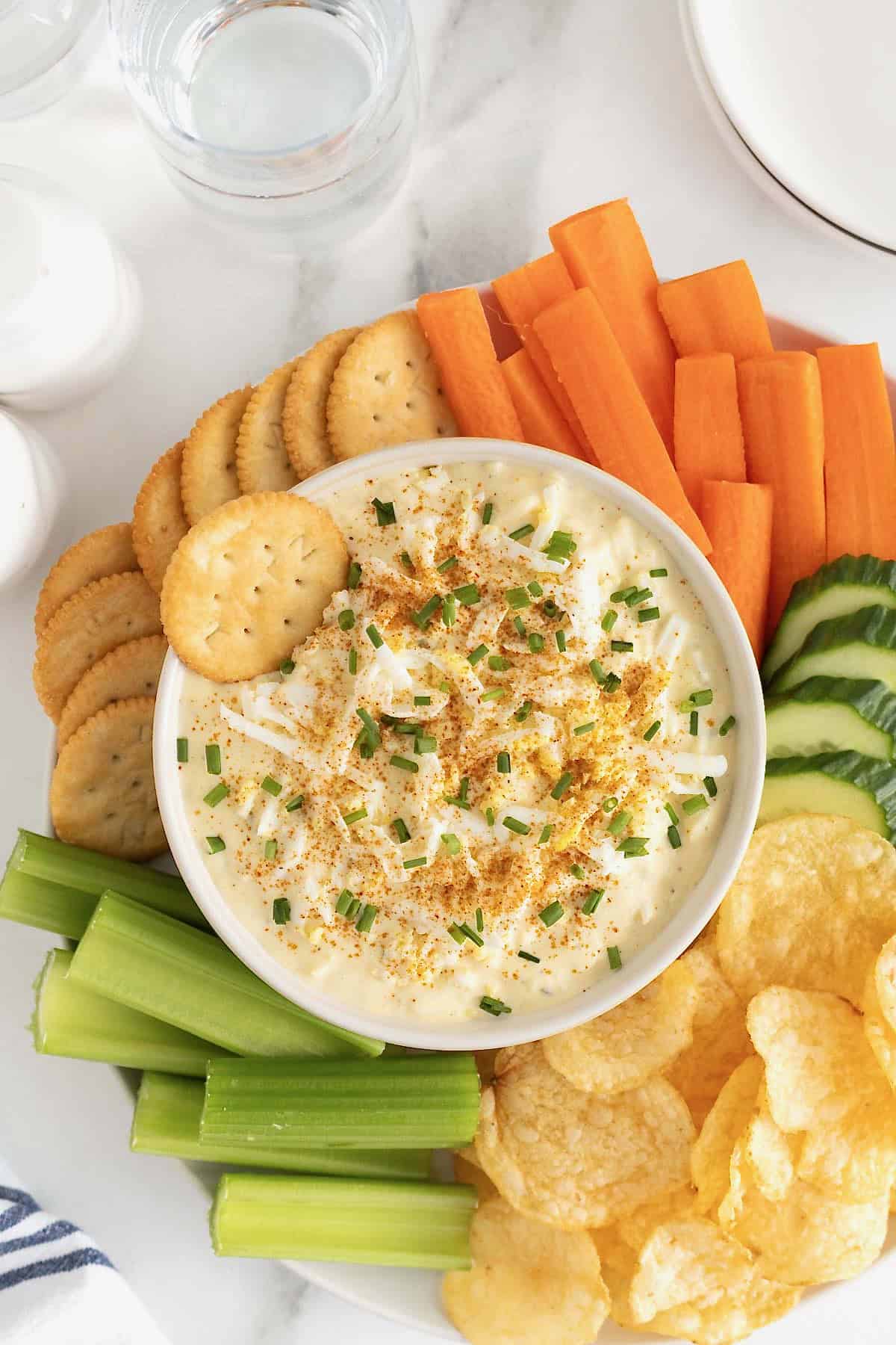 A large white bowl of deviled egg dip surrounded by crackers, carrot sticks, cucumber slices, potato chips and celery sticks on a large round platter.