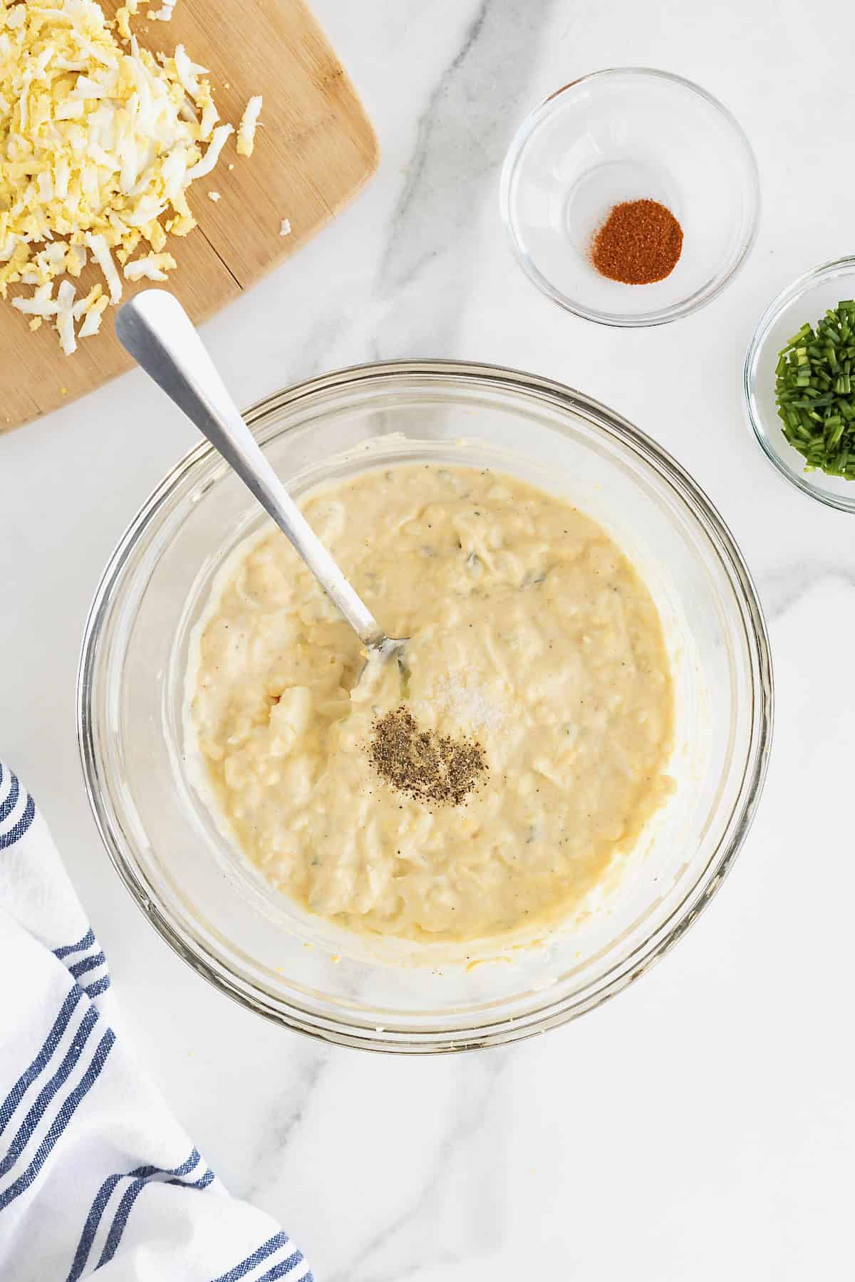 Salt and pepper being added to a large glass mixing bowl of deviled egg dip.