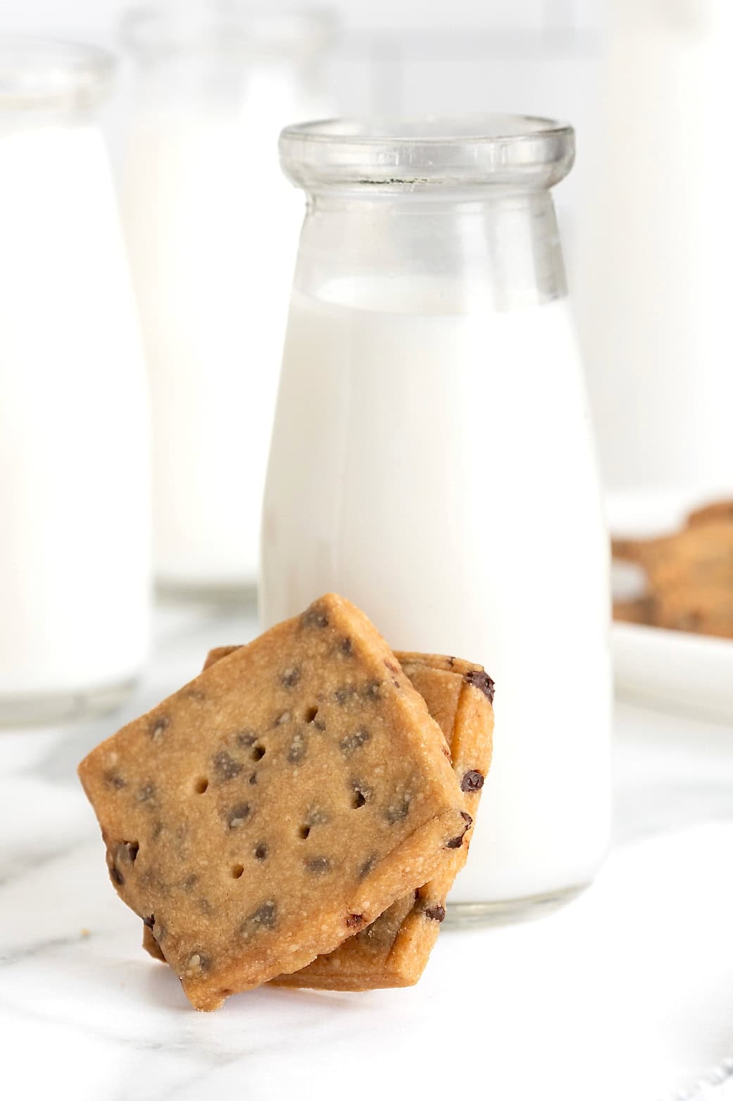 Two chocolate chip cookie crisps leaning against a glass carafe of milk.
