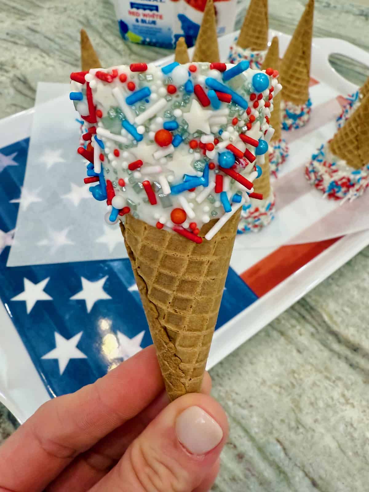 A sugar cone with the top coated in vanilla candy coating and red, white and blue sprinkles.